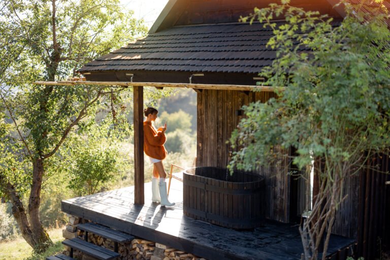 A woman in a cozy sweater enjoys her morning coffee on a wooden porch with an outdoor bath, surrounded by greenery and sunlight, embracing the tranquility of nature